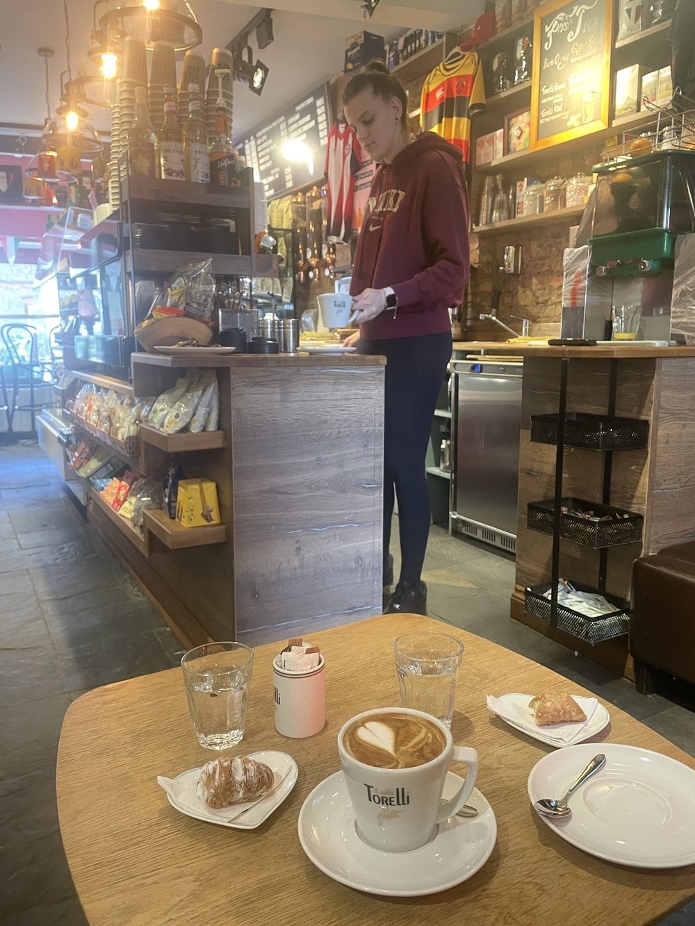 Inside Café Torelli looking toward the counter and deli cabinet, with a cappuccino and pastries on a wooden table in the foreground
